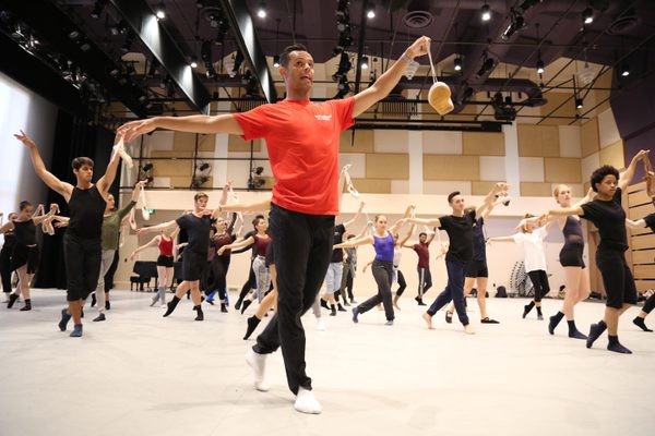 Dance class in studio led by man in red top and black trousers reaches one arm forward holding a shoe by its lace