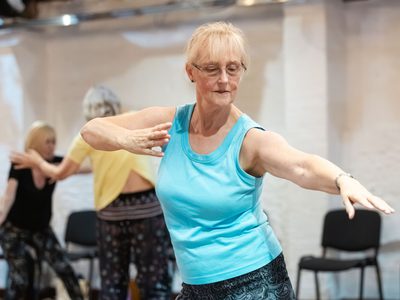 A person in a blue t-shirt and glasses is participating in a dance class. She is focussed on the movement and has one arm out to her side and the other bent towards her chest.