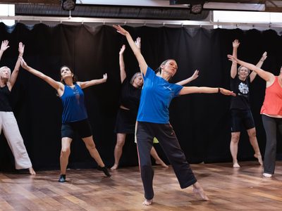An instructor stand at the front of a dance class demonstrating a movement with her arms reaching out to her sides, the participants imitate the same movement.