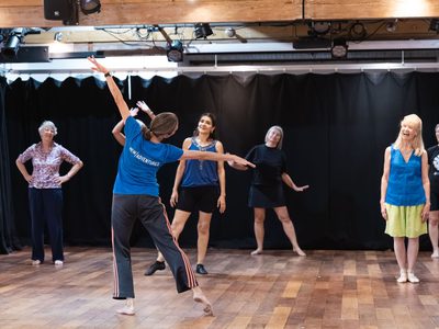 An instructor in a blue t-shirt is demonstrating a movement in a dance class. She is reaching her arms out at her sides, and the participants watch.