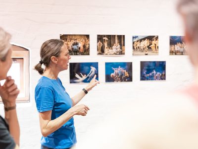 An instructor in a blue t-shirt is pointing to images of a dance production which are stuck to a wall. People stand around listening intently.