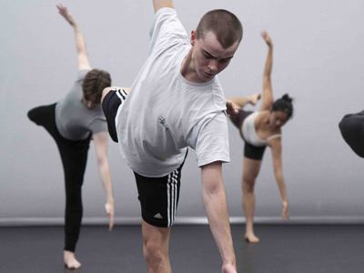 People are dancing in a dance studio. The young person in the foreground is leaning forward, balancing on one leg with the other leg stretched behind them. They have one arm reaching upwards and one reaching towards the floor.