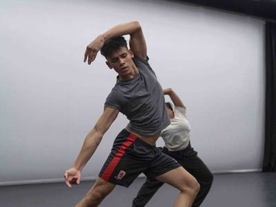 A young man is dancing. His front leg is bent at the knee and the other is straight and stretched out behind him. His back is slightly arched with one arm reached over his head and the other following the line of his back leg.