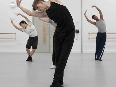 People are dancing in a studio. The young man in the foreground is bending over to one side, his arms arched about his head. The other people in the room are carrying out the same movement.